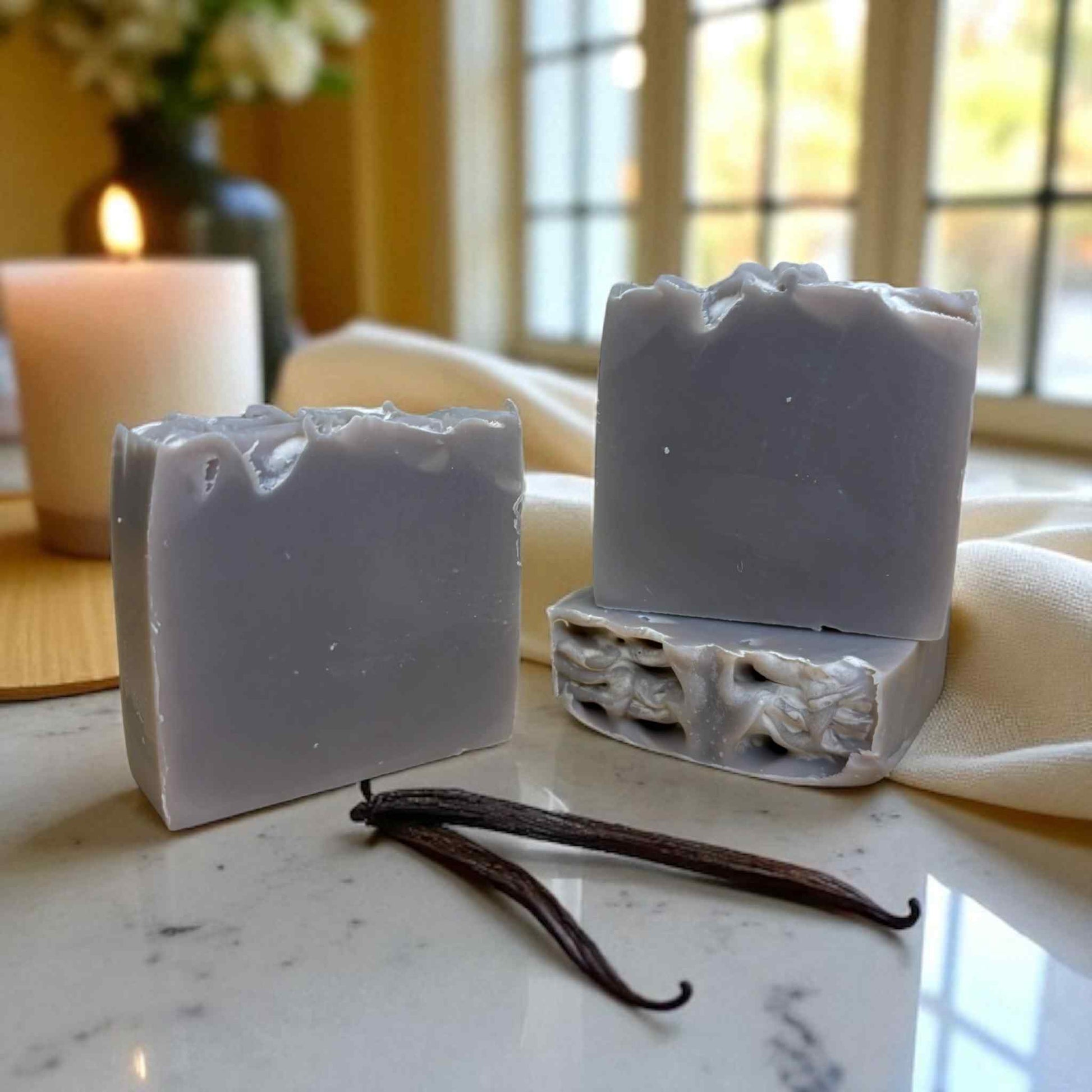 Two bars of gray soap on a marble surface with vanilla beans and a blurred background.