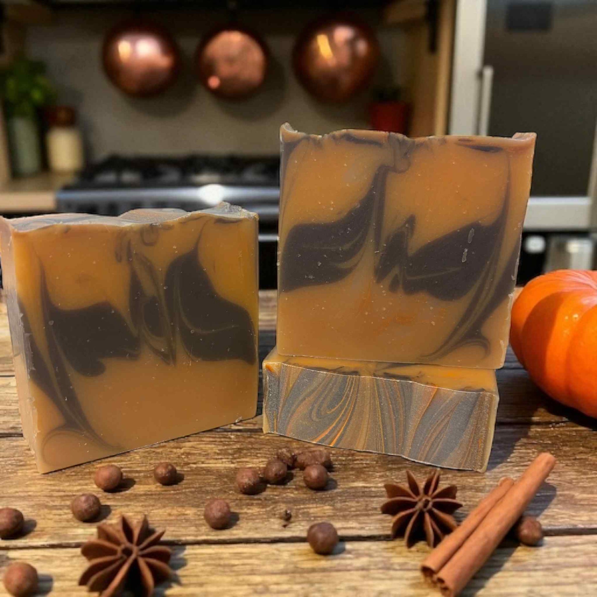 Two swirled soap bars on a wooden surface with spices and a pumpkin in the background.