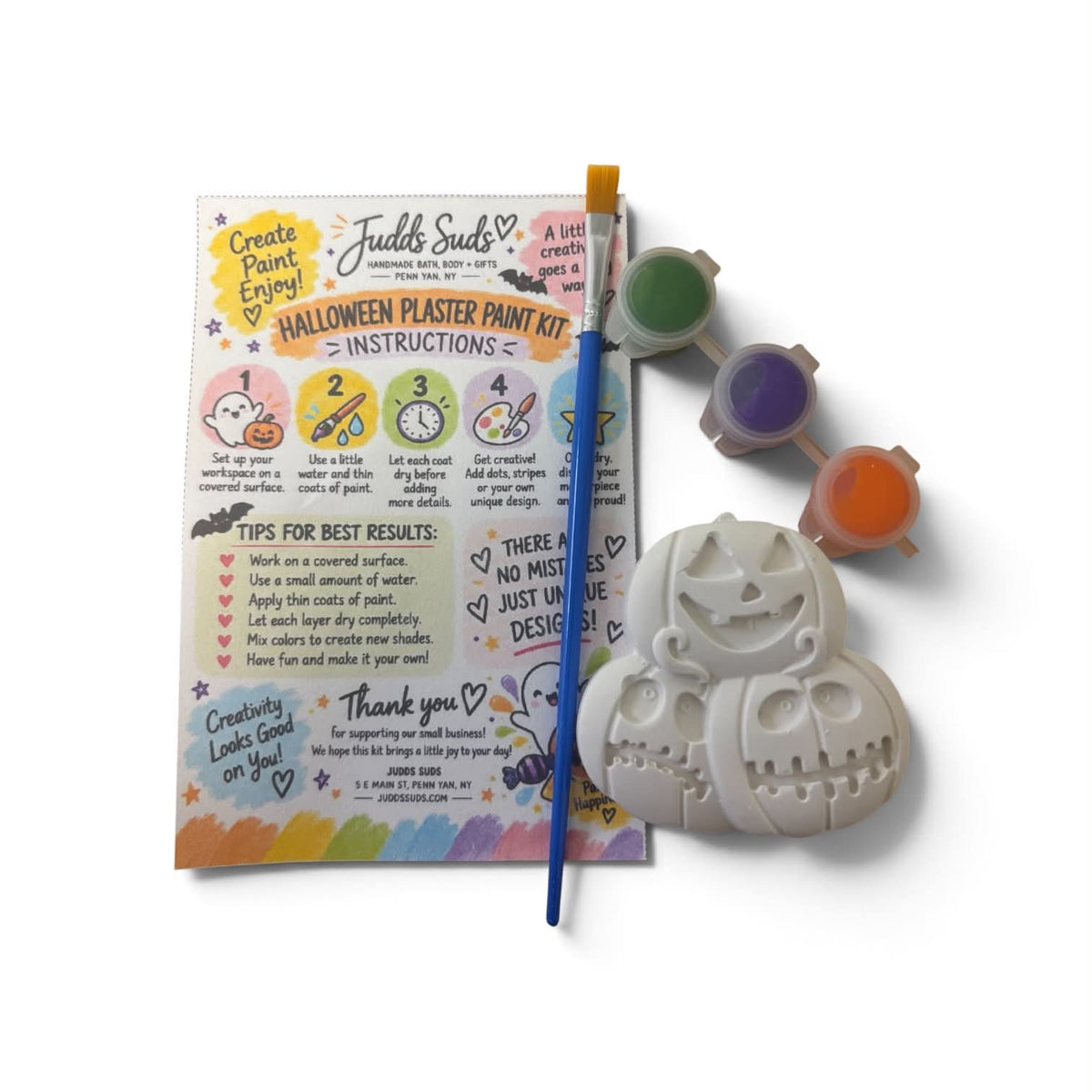 Halloween Plaster Paint Kit by Judd Seeds, featuring a pumpkin-shaped plaster mold, a paintbrush, three small paint wells with green, purple, and orange paint, and an instruction booklet on a white background.